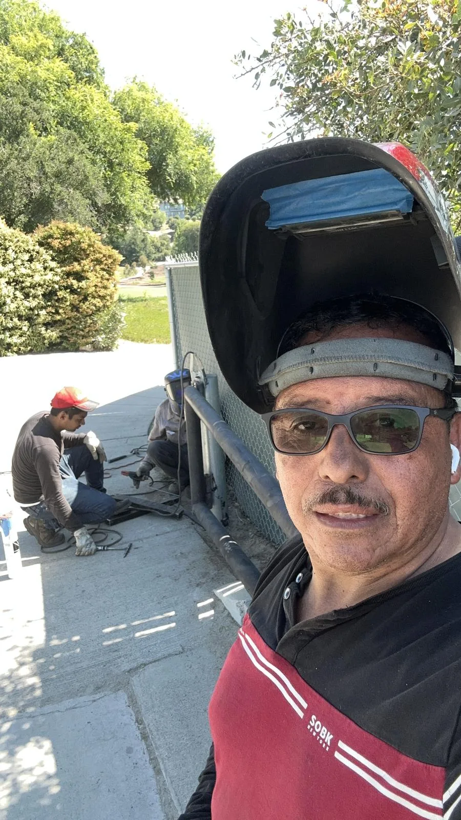 A worker wearing a welding helmet with other workers beside a metal structure outdoors